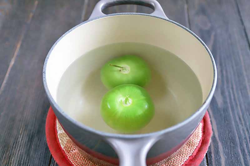 Two green tomatillos blanching in a blue and cream-colored enameled saucepan of hot water on a beige and red round potholder, on a dark brown wood surface.