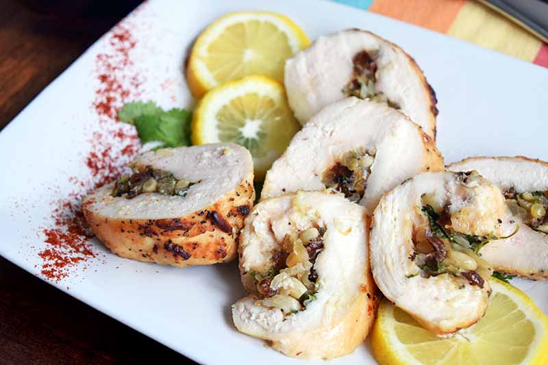 Horizontal image of chicken roulade stuffed with nuts, raisins, and herbs, on a white serving platter with thin lemon slices, a sprig of parsley, and a sprinkle of paprika, on a brown wood surface with a multicolored cloth napkin topped with silverware in the top left corner of the frame.