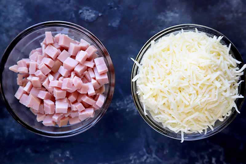 Horizontal overhead photo of two glass bowls of chopped cooked ham and grated white cheddar cheese, on a dark blue background with white splotches.