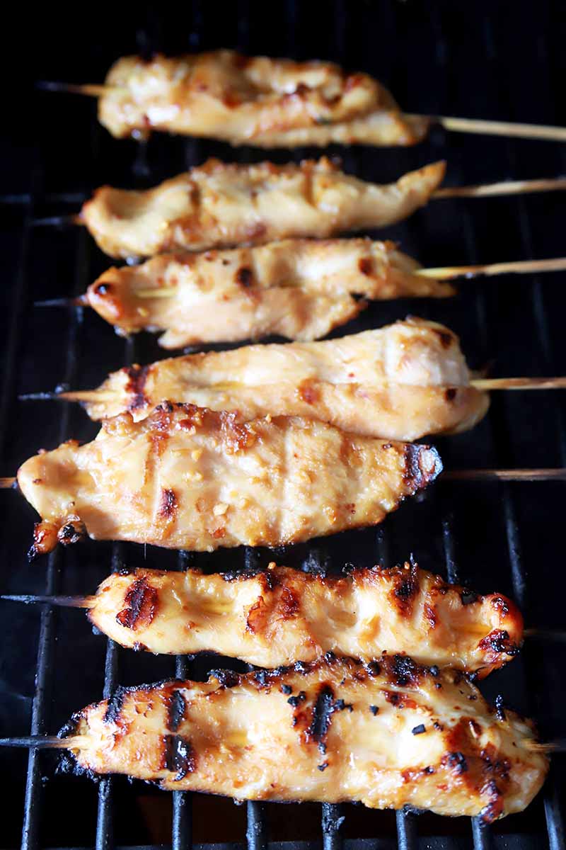 Vertical overhead image of seven bamboo skewers of marinated chicken being cooked on a hot grill, with a black background.