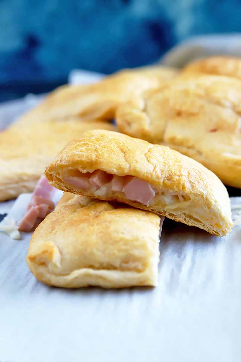 Vertical head-on shot of a savory ham and cheese pastry that has been cut in half to show the filling inside and stacked, on a white piece of parchment paper, with more of the snacks in the background, against a dark and light blue mottled backdrop.