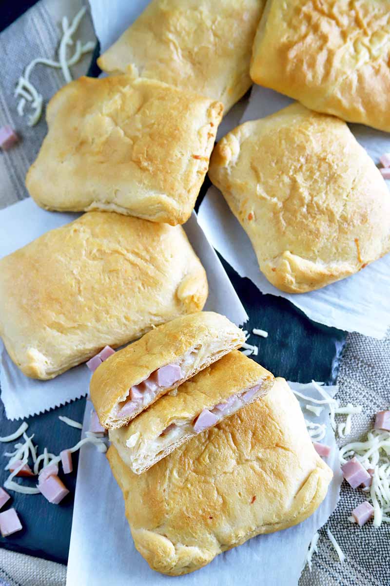 Vertical overhead image of savory ham and cheese pastries, with one at the bottom of the frame cut in half to show the filling inside and stacked on another piece, on squares of white parchment paper on top of a dark gray slate surface, with scattered grated cheese.