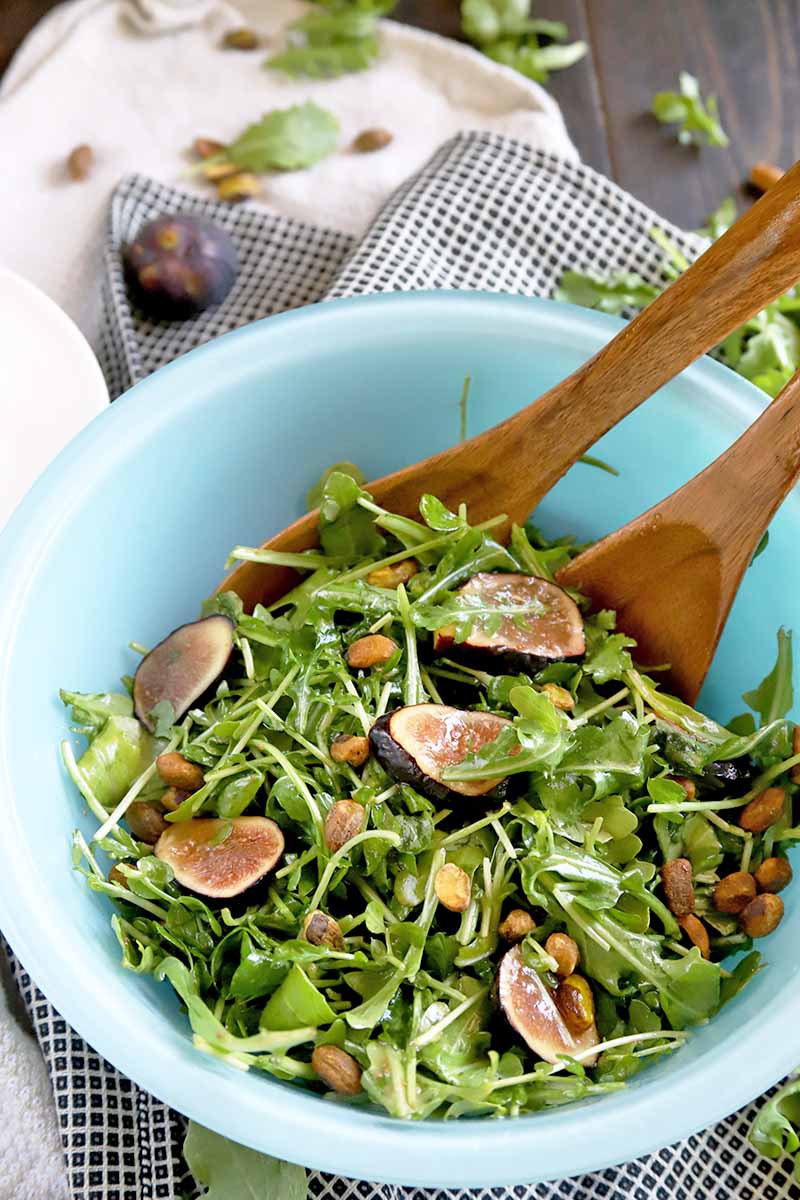 Oblique overhead vertical image of a blue glass bowl of salad made with greens, nuts, and fresh figs, with wooden serving utensils, on a brown wood surface with scattered pistachios, fruit, and leafy greens, on a folded cloth.