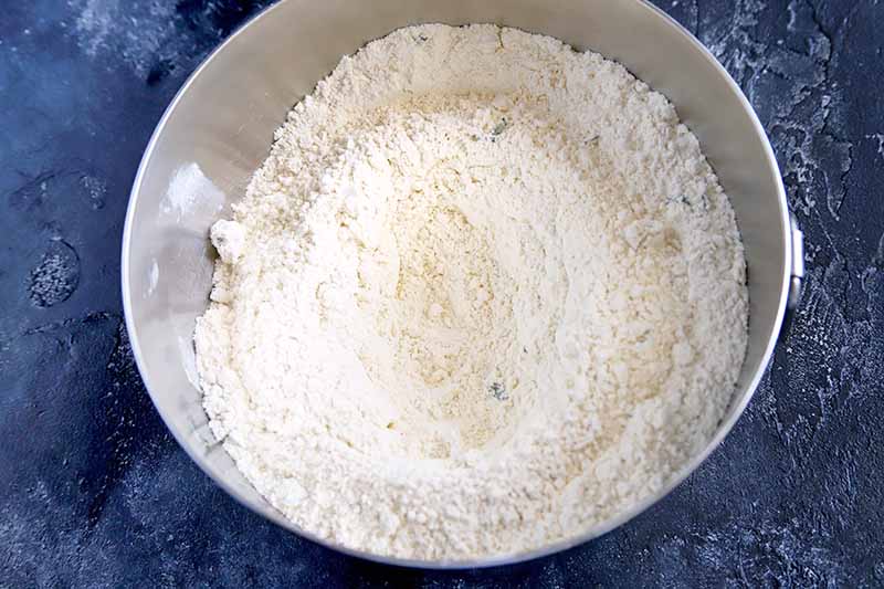 Overhead horizontal shot of a dry flour mixture in a stainless steel bowl, on a navy blue surface.