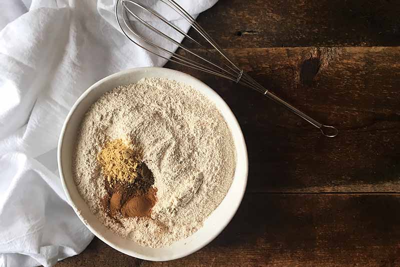 Horizontal image of a bowl of flour with piles of spices next to a whisk.