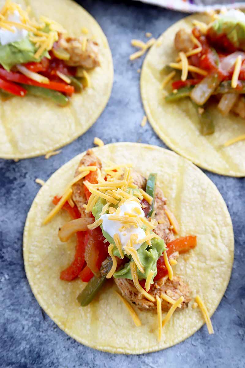 Vertical image of three soft-shell tacos topped with chicken, green and red peppers, and various condiments.