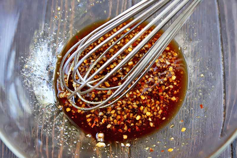 Overhead closeup horizontal image of a wire whisk stirring a marinade of minced garlic, ginger, soy sauce, and red chili flakes in a glass mixing bowl, on a grayish brown wood surface.