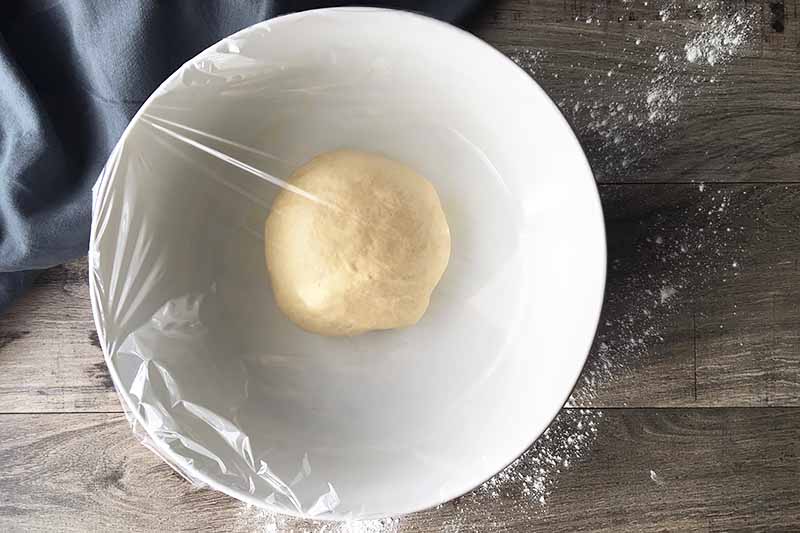 Horizontal image of a small mound of dough in a white bowl covered in plastic wrap.