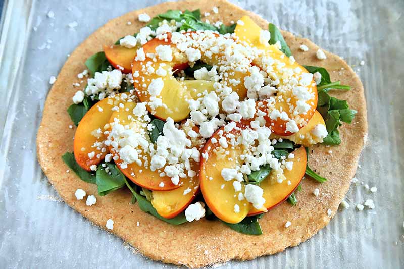 Horizontal image of rolled out pizza dough topped with baby spinach leaves, sliced peaches, and crumbled goat cheese, on a white paper surface.