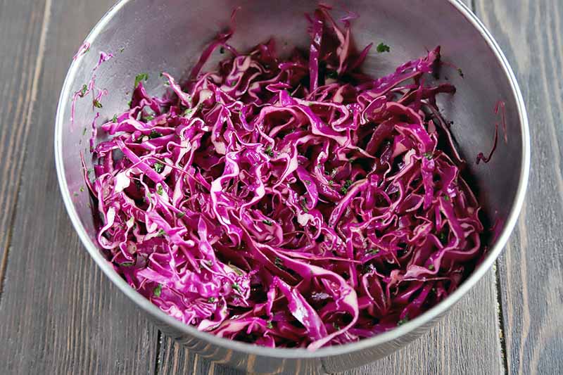 A stainless steel bowlful of red shredded cabbage slaw, on a dark brown wood surface.
