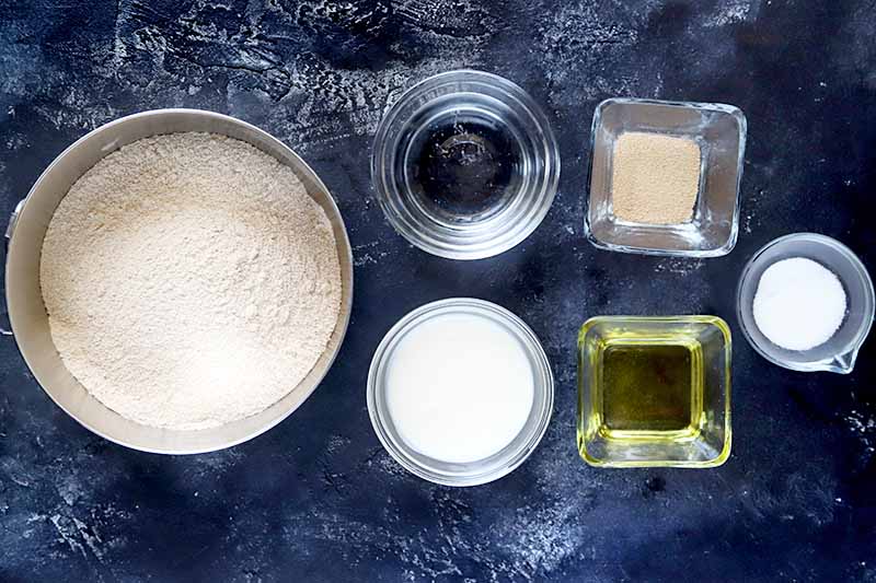 Overhead horizontal image of medium-sized stainless steel mixing bowl of spelt flour to the left of two small round glass bowls of kefir and water, two small square glass dishes of dry yeast and olive oil, and a very small bowl of salt, on a dark blue surface with white streaks.