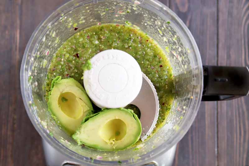 Horizontal overhead image of a food processor filled with a tomatillo sauce and two halves of a peeled ripe avocado, on a dark brown wood surface.