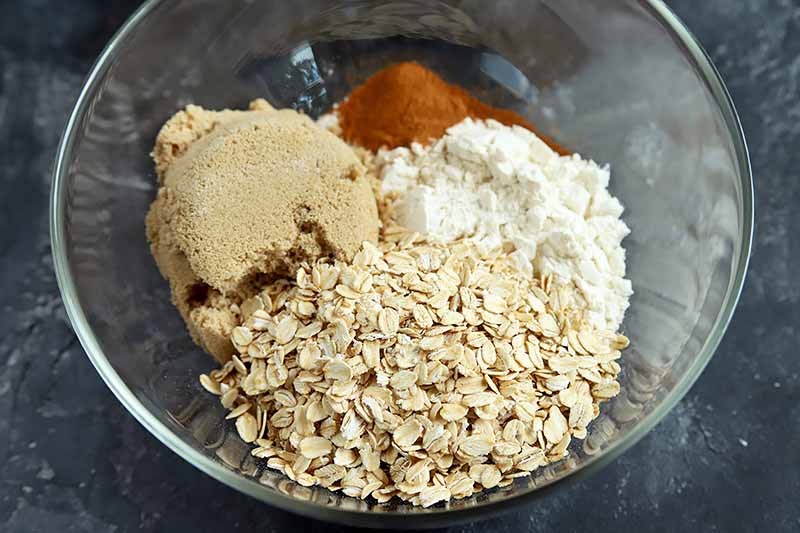 Horizontal overhead closely cropped image of a bowl containing separate piles of ground cinnamon, packed light brown sugar, rolled oats, and flour, on a gray surface with white streaks.