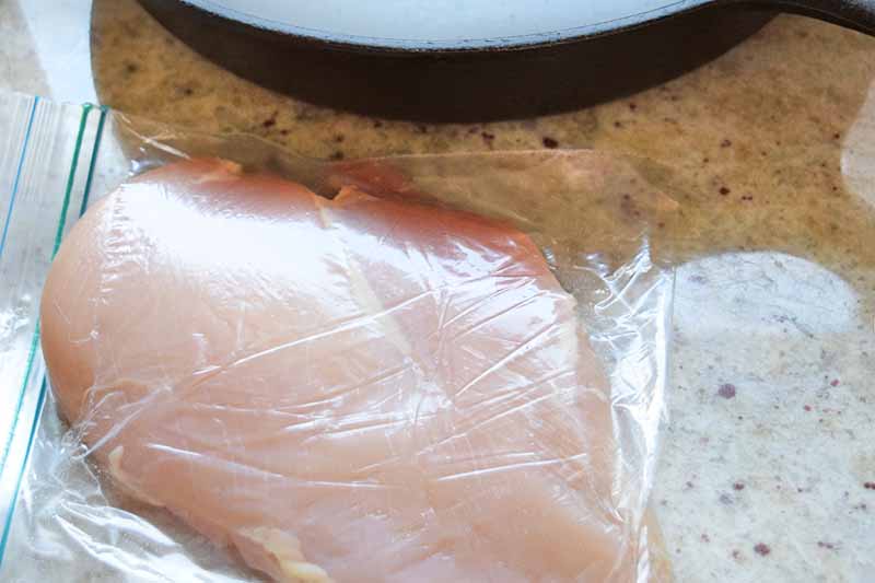Horizontal overhead closeup image of a thinly pounded chicken breast in a zip-top plastic bag beside a cast iron pan, on a beige speckled countertop.