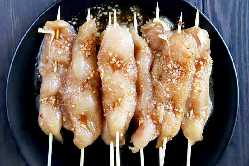 Horizontal overhead image of a black plate with bamboo skewers of marinated raw chicken on top, on a grayish brown wood surface.