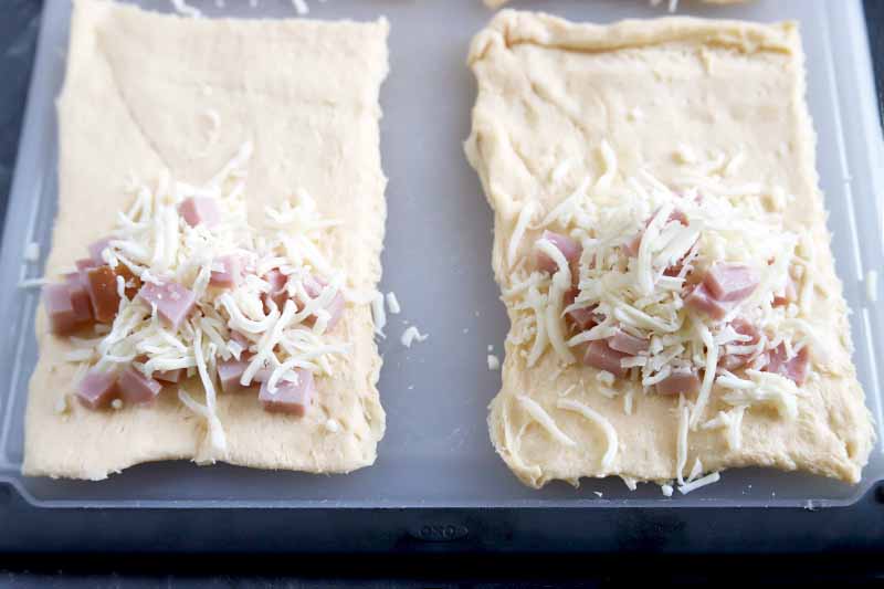 Horizontal oblique overhead image of rectangles of rolled out dough on a white plastic cutting board with a black border, with small piles of diced ham and shredded cheese on half of the dough.