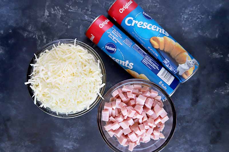 Horizontal overhead image of two cylindrical cans of PIllsbury crescent roll dough beside two glass bowls of shredded white cheddar cheese and diced cooked ham, on a gray surface.