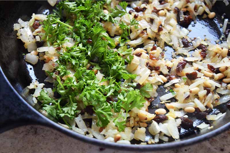 Closely cropped horizontal overhead image of sauteed onion and garlic with toasted pine nuts and chopped fresh parsley in a cast iron pan.