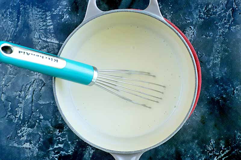 Horizontal overhead image of a gray and cream-colored saucepan resting on a patterned dark blue surface, with a wire whisk with a sky blue handle stirring a thick white liquid.