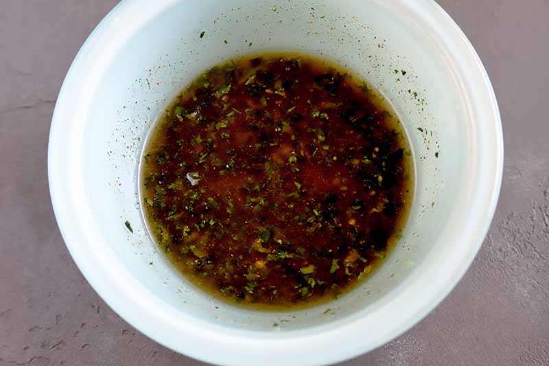 Horizontal overhead image of a white bowl of marinade made with oil, lime juice, and spices, on a gray surface.