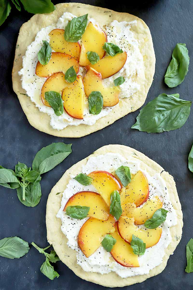 Vertical overhead image of two round flatbreads topped with ricotta, thinly sliced peaches, and basil leaves, with basil sprigs and leaves scattered around them on a dark blue surface.