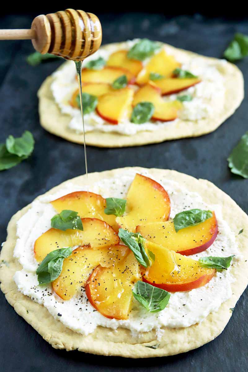 Vertical image of a wooden dipper drizzling honey onto a round peach, ricotta, and basil flatbread, with another identical appetizer in soft focus in the background, on a dark gray surface with scattered fresh basil leaves, against a black backdrop.
