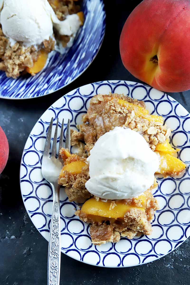 Vertical overhead closely cropped image of a patterned blue and white plate of peach crisp topped with vanilla ice cream with a fork, with another plate of the same at the top left corner of the frame and two whole stone fruits with orange and white skins, on a gray surface.