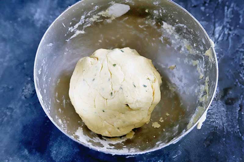 A ball of dough with flecks of green herbs rests in the bottom of a stainless steel bowl that is also streaked with dough, on a dark gray surface.