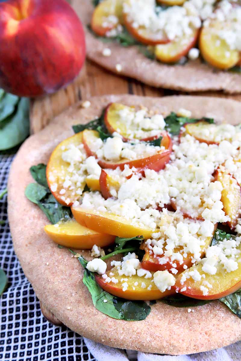 Vertical closely cropped image of a peach, spinach, and goat cheese pizza, with another in the background, with a whole stone fruit and leafy greens on a black and white checkered cloth.