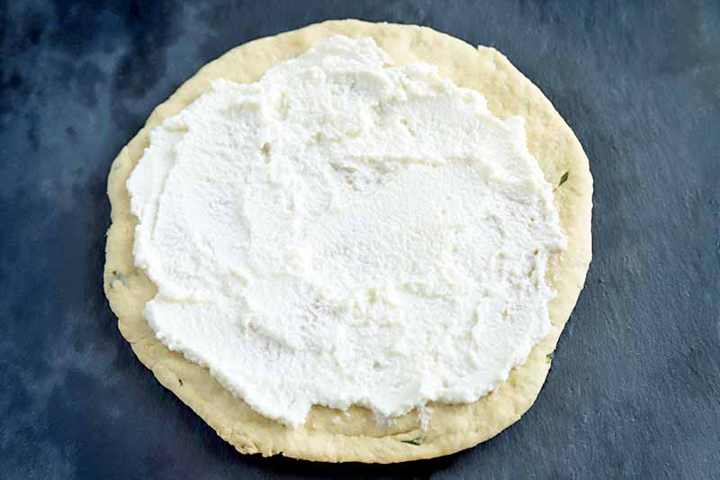 Horizontal image of a round baked dough circle with a thin layer of ricotta cheese spread on top, on a dark grayish blue surface.