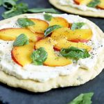 Horizontal image of a round flatbread topped with a thin layer of ricotta cheese, thinly sliced peaches, and basil leaves, on a gray slate surface with another identical appetizer in the background and scattered herbs.