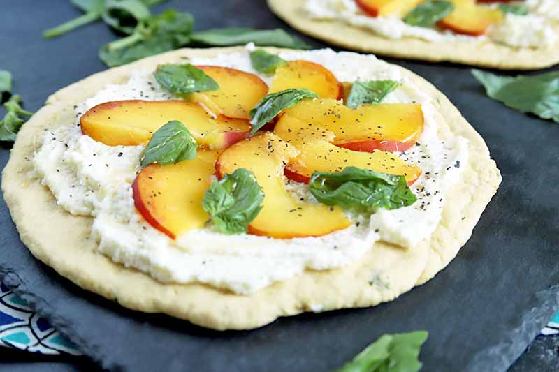 Horizontal image of a round flatbread topped with a thin layer of ricotta cheese, thinly sliced peaches, and basil leaves, on a gray slate surface with another identical appetizer in the background and scattered herbs.