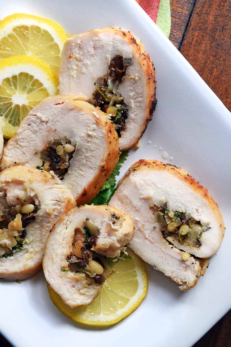 Vertical overhead closely cropped image of homemade chicken roulade on a white rectangular ceramic serving platter with thinly sliced lemon, on a brown wood surface.