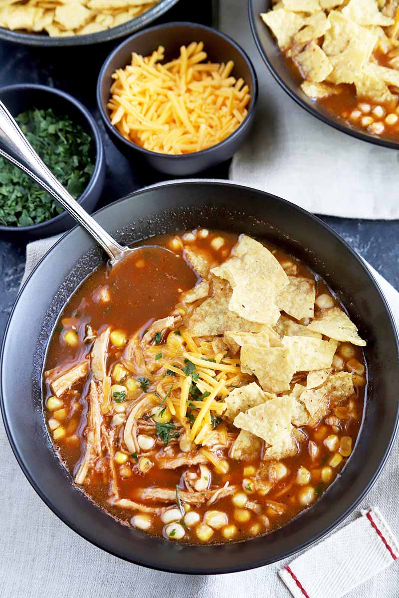 Vertical overhead image of a black ceramic bowl of barbecue chicken soup with a spoon in it and crushed tortilla chips on top, with another identical bowl in the top right corner of the frame, and three smaller bowls of chips, chopped green herbs, and shredded orange cheddar cheese at the top right, on a striped white cloth on top of a gray surface.