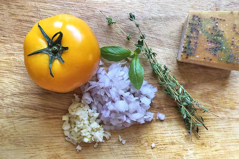 Horizontal overhead image of a golden tomato with a green top, minced garlic and shallow, a few sprigs of thyme, and a hunk of Parmesan cheese with a yellowish rind, on a blonde wood cutting board.