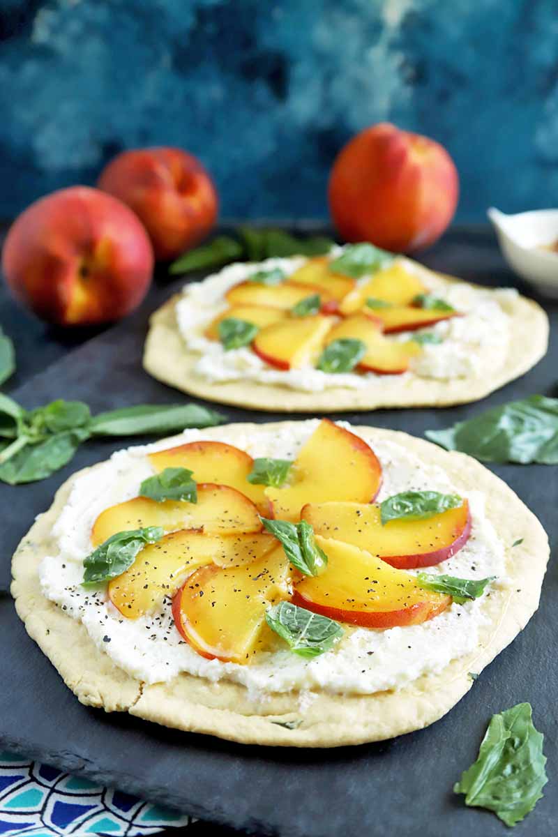 Vertical image of two round flatbreads topped with ricotta, sliced peaches, and basil, on a slate surface with scattered herb leaves and three whole peaches in the background, on a dark and light blue patterned cloth, against a black and blue mottled background.
