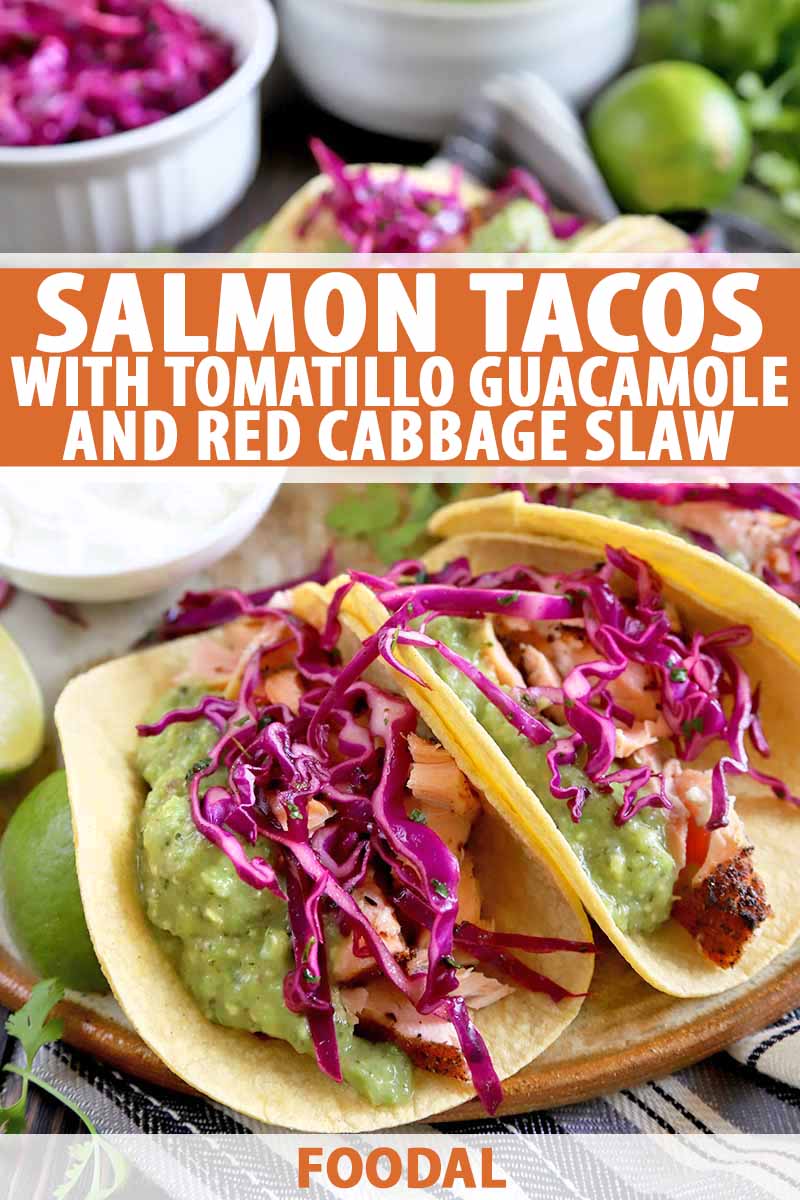 Vertical image of salmon tacos in yellow corn tortillas, with tomatillo guacamole and red cabbage slaw on top, on a brown and beige serving platter with limes, and small dishes of slaw and sour cream, on a brown wood table, printed with orange and white text near the midpoint and at the bottom of the frame.