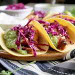 Horizontal image of several salmon tacos in corn tortillas on a tan and brown ceramic serving platter, topped with homemade tomatillo guacamole and red cabbage slaw, on a striped blue, gray, and white cloth with scattered sprigs of cilantro, with small dishes of slaw, sauce, and sour cream in the background, on a brown wood surface.