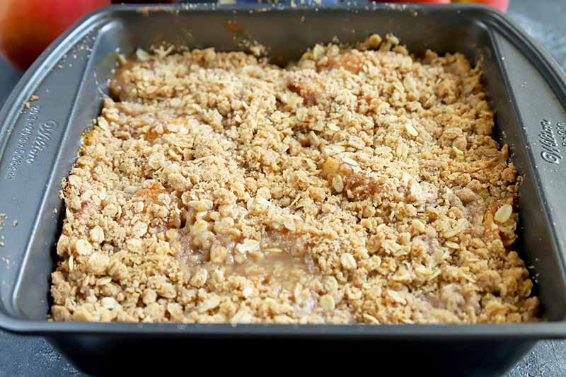 Horizontal image of a metal baking pan filed with a brown sugar and cinnamon crumb mixture, on a gray surface with a peach in soft focus in the background.