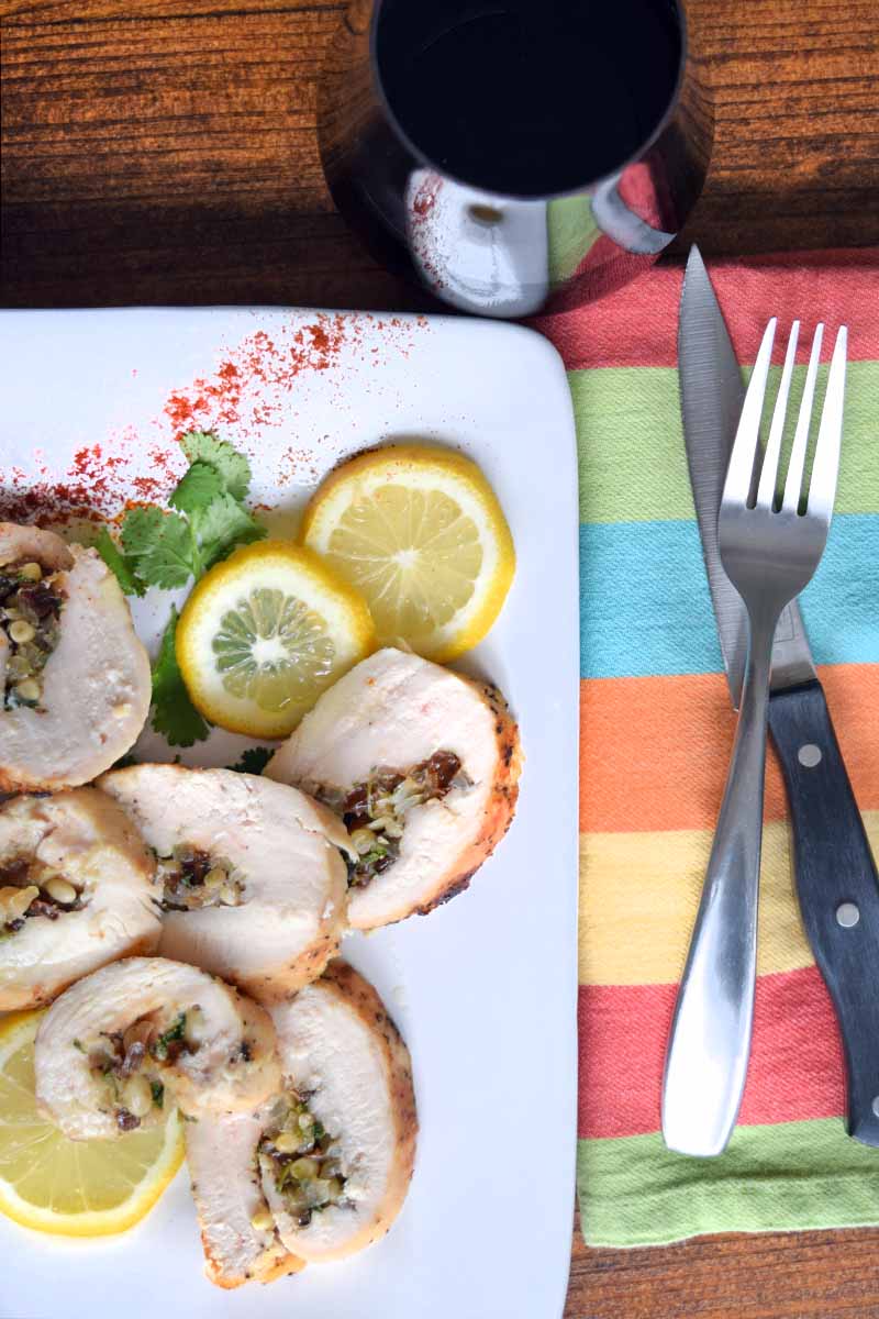 Vertical overhead image of a white ceramic serving platter of chicken roulade with slices of lemon, a sprig of fresh herbs, and a sprinkle of paprika, on a brown wood table with a glass of red wine and a striped multicolored cloth napkin topped with a fork and a steak knife.