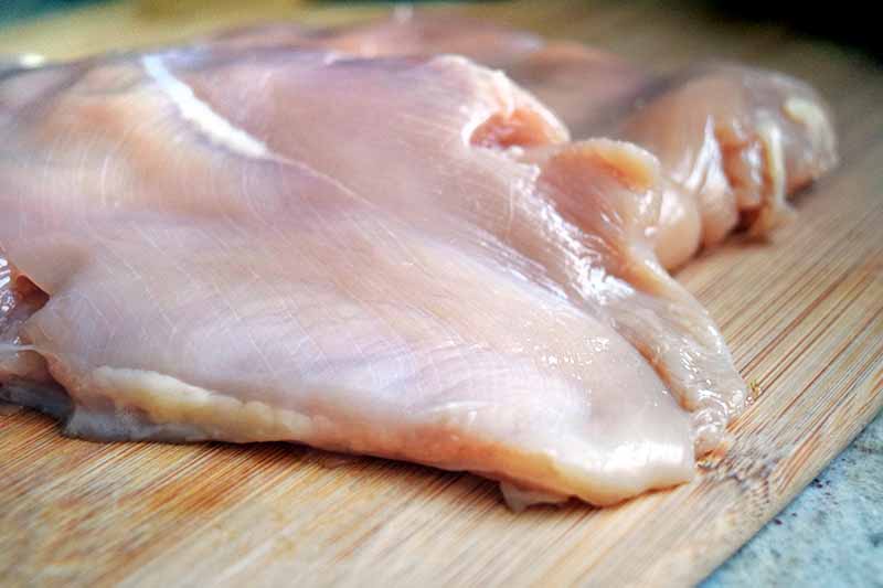 Horizontal image of a boneless skinless chicken breast that has been pounded flat, on a wooden cutting board on top of a gray speckled countertop.