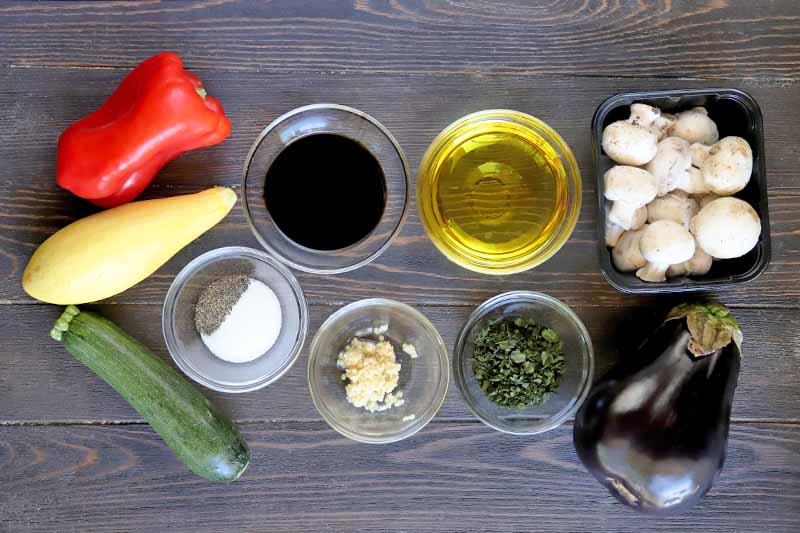 Horizontal image of fresh vegetables, oil, garlic, vinegar, and flavorings in small bowls.