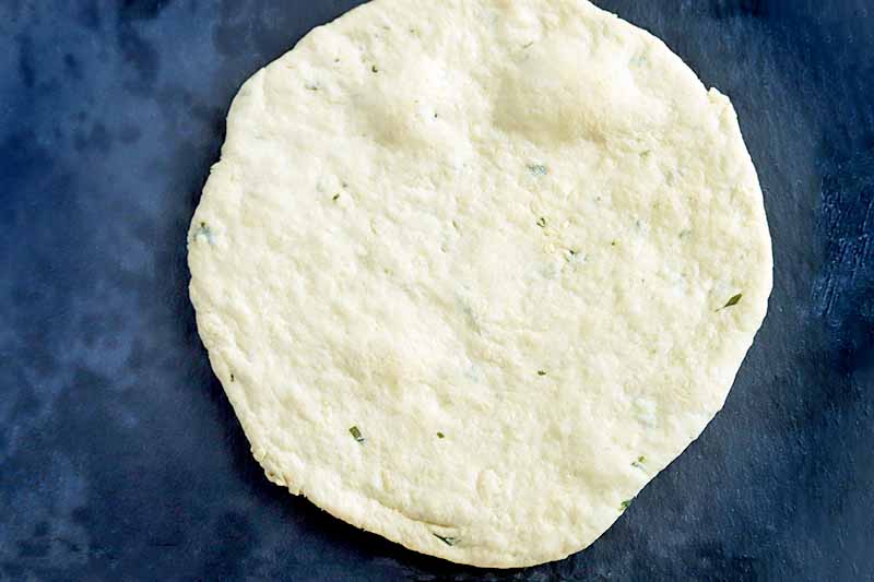 Overhead horizontal image of a freshly baked round of herbed flatbread dough, on a dark gray surface.
