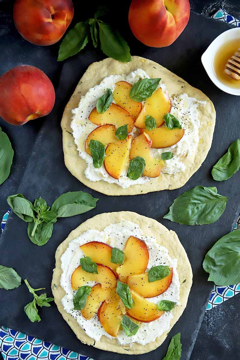 Vertical overhead image of two round peach, basil, and ricotta flatbreads on a gray slate surface, with three whole peaches, scattered basil leaves, two dark and light blue patterned cloths, and a small white bowl of honey with a wooden dipper.