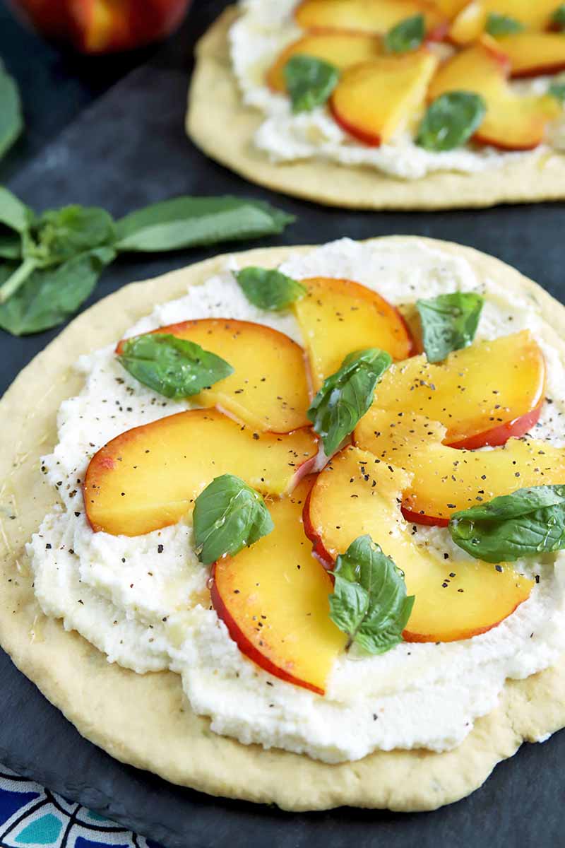 Closeup vertical image of a peach, basil, and ricotta flatbread on a slate surface with another in soft focus in the background, with scattered basil leaves,.