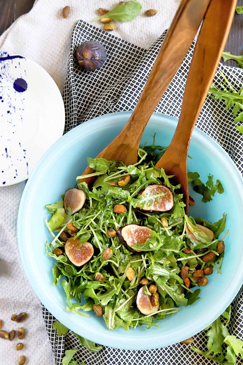 Vertical overhead image of a light blue glass bowl of arugula with sliced figs and pistachios, with wooden serving utensils, on a dark brown wood table topped with a gathered white cloth, with a blue and white plate and scattered nuts, fruit, and leaves.