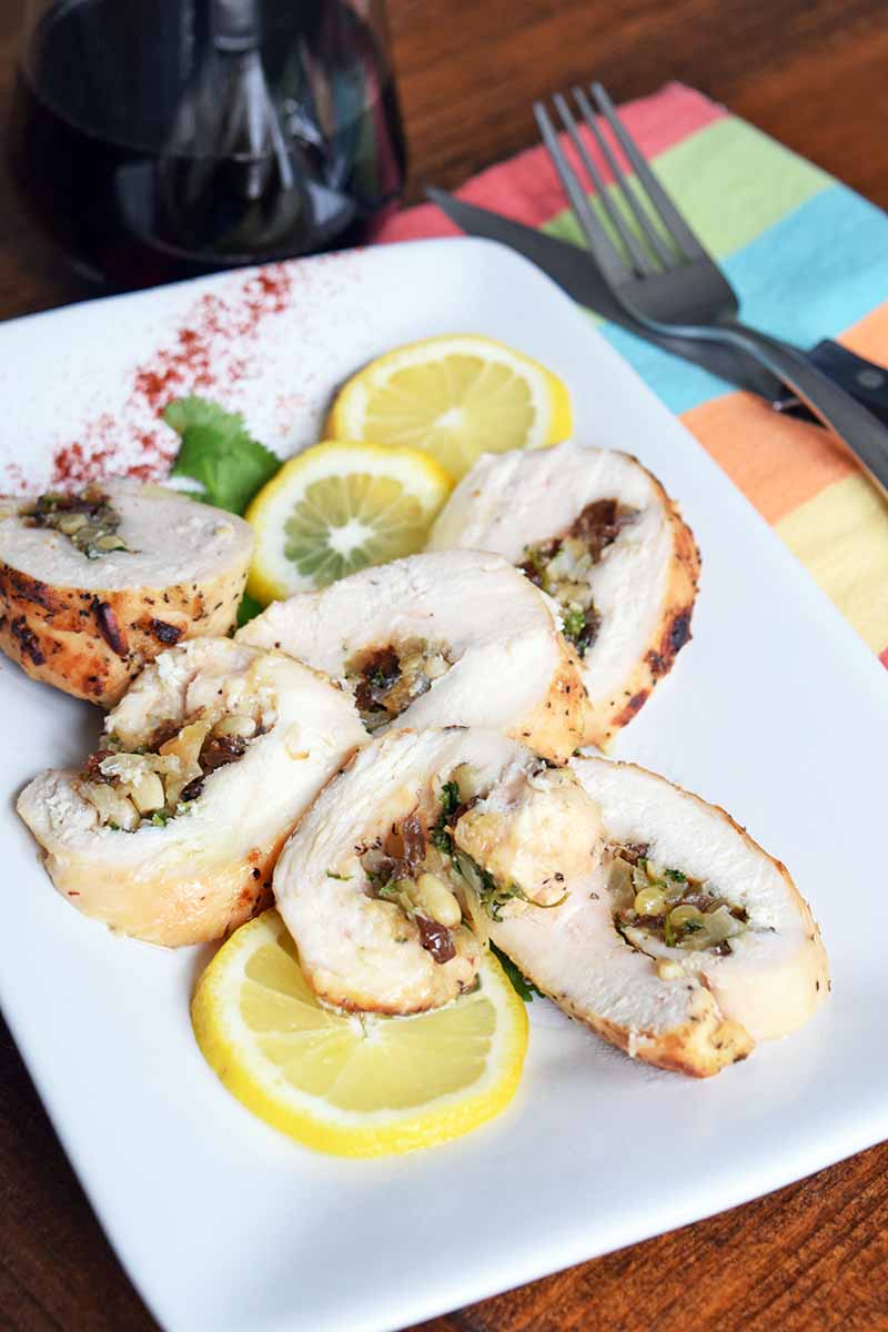 Horizontal oblique overhead image of a white serving platter of homemade chicken roulade slices with thin lemon slices, a sprig of fresh herbs, and a sprinkle of paprika for garnish, on a brown wood table with a stemless glass of red wine, and a folded striped multicolored cloth napkin topped with a knife and fork.
