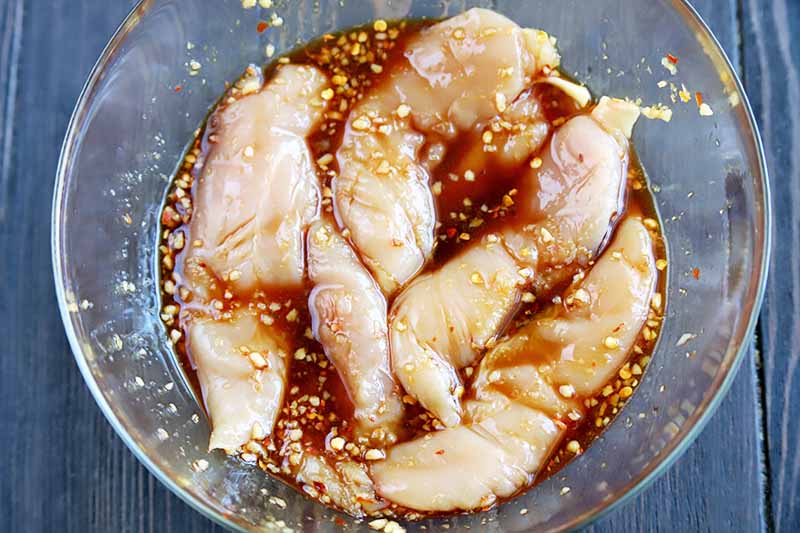 Horizontal overhead closely cropped image of raw chicken tenderloins in a glass bowl, on a marinade of soy sauce, ginger, and garlic, on a gray surface.