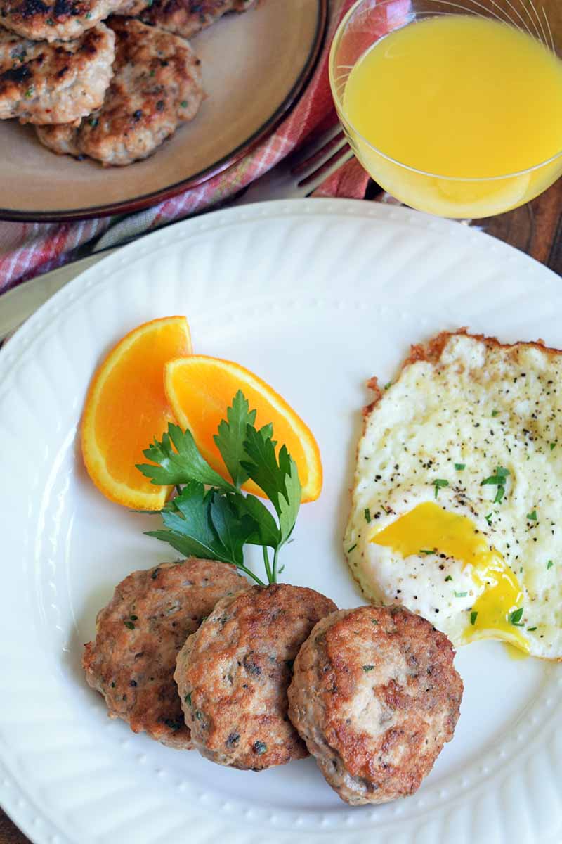 Vertical overhead image of a white ceramic plate with three round turkey sausage patties arranged in a row, two orange wedges, a sprig of flat leaf parsley, and a fried egg with a broken yolk oozing out, a glass of orange juice, and a platter of more of the breakfast meat at the top left of the frame.
