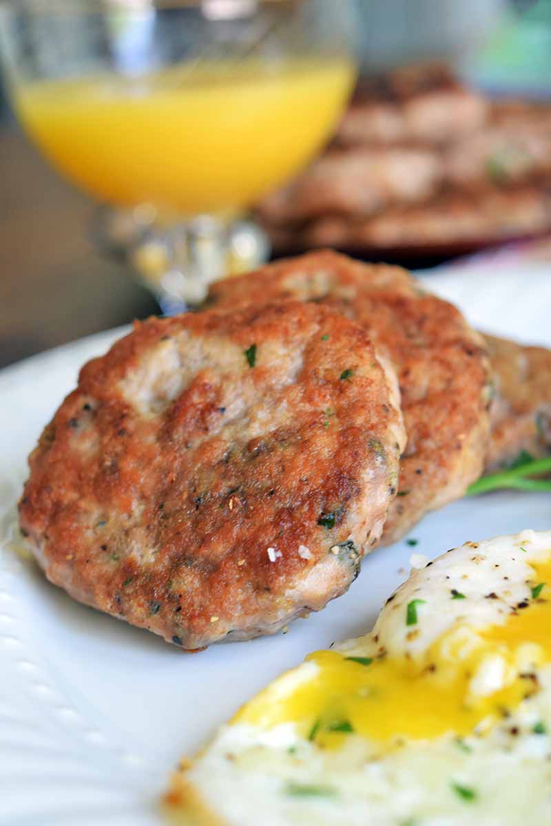 Vertical image of three fried turkey sausage patties on a white plate with a fried egg that has been broken to show the liquid yolk inside, with a glass of of orange juice and a platter of more of the homemade breakfast meat in soft focus in the background.